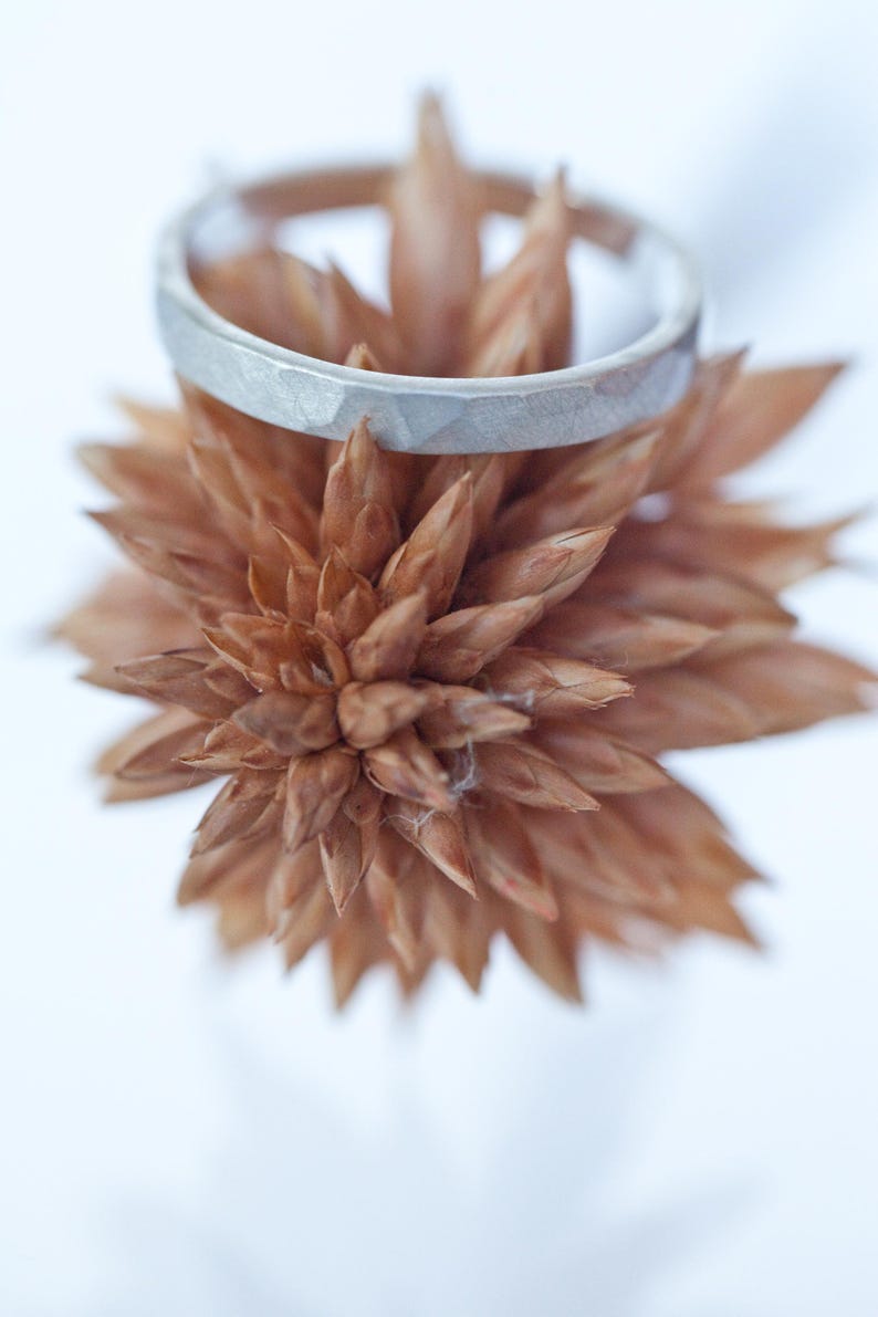 Może przedstawiać: Srebrny pierścionek o teksturowanej powierzchni spoczywa na wysuszonym, brązowym kwiatku. Pierścionek ma geometryczny wz&oacute;r i matowe wykończenie. Kwiat ma wiele spiczastych płatk&oacute;w, tworząc naturalną estetykę. Tło jest miękkie, rozmyte białe.