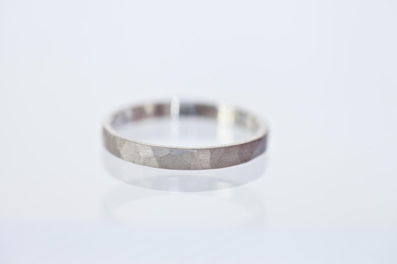 Może przedstawiać: Srebrny pierścionek o młotkowanej fakturze. Pierścionek ma prosty kształt obrączki, z lekko nieregularną powierzchnią. Pierścionek jest prezentowany na białej powierzchni, odbijając światło.