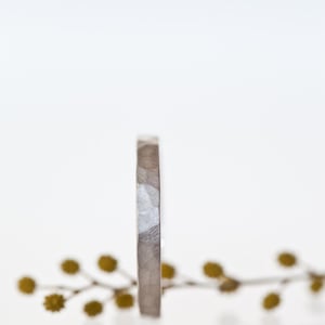 Może przedstawiać: Srebrny pierścionek o teksturowanej powierzchni stoi na białym tle. Pierścionek ma prosty, nowoczesny design z lekko młotkowanym wyglądem. Gałązka ż&oacute;łto-zielonych kwiat&oacute;w znajduje się w tle.