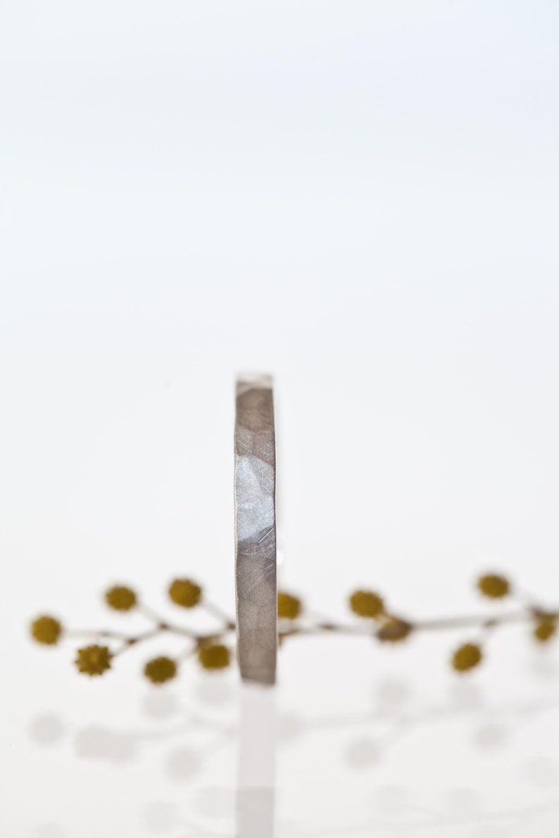 Może przedstawiać: Srebrny pierścionek o teksturowanej powierzchni stoi na białym tle. Pierścionek ma prosty, nowoczesny design z lekko młotkowanym wyglądem. Gałązka ż&oacute;łto-zielonych kwiat&oacute;w znajduje się w tle.