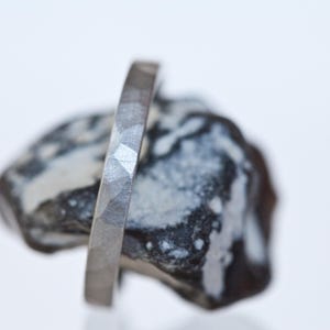 Może przedstawiać: Prezentowany jest srebrny pierścionek o fasetowanej powierzchni. Pierścionek jest osadzony na teksturowanym, ciemnoszarym i białym kamieniu. Projekt pierścionka charakteryzuje się serią małych, kątowych cięć, tworzących unikalny, teksturowany wygląd. Pierścionek to biżuteria.