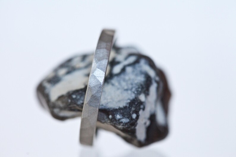 Może przedstawiać: Prezentowany jest srebrny pierścionek o fasetowanej powierzchni. Pierścionek jest osadzony na teksturowanym, ciemnoszarym i białym kamieniu. Projekt pierścionka charakteryzuje się serią małych, kątowych cięć, tworzących unikalny, teksturowany wygląd. Pierścionek to biżuteria.