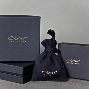 Może przedstawiać: Asortyment czarnych pudełek na biżuterię i woreczek ze sznurkiem. Pudełka są prostokątne i ułożone w stos, ze złotymi literami z napisem "Cirer sea jewelry". Woreczek r&oacute;wnież ma złote litery.