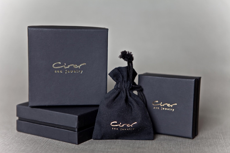 Może przedstawiać: Asortyment czarnych pudełek na biżuterię i woreczek ze sznurkiem. Pudełka są prostokątne i ułożone w stos, ze złotymi literami z napisem "Cirer sea jewelry". Woreczek r&oacute;wnież ma złote litery.