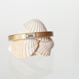 May include: A gold-colored ring with a rectangular, clear gemstone set in the center. The ring rests on a collection of beige seashells, set against a white background. The ring's band has a matte finish.
