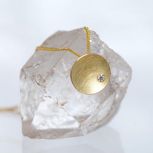 May include: A gold pendant necklace featuring a brushed, circular pendant with a small, sparkling gemstone. The necklace is displayed on a clear, rough-cut crystal, creating a contrast of textures and colors.