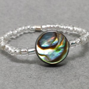Puede incluir: Anillo de cuentas con una pieza central redonda de concha de abulón iridiscente. La banda del anillo está hecha de pequeñas cuentas transparentes. La concha de abulón tiene una mezcla de colores verdes, azules y dorados.
