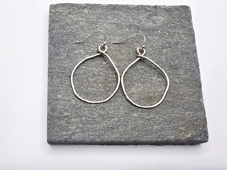 May include: A pair of silver-coloured, open-hoop earrings. The earrings have a hammered texture and a teardrop shape. They are displayed on a grey, textured surface.