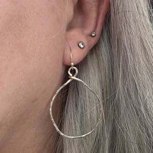 May include: Close-up of a person's ear with silver hoop earrings. The earrings are large, hammered metal hoops with a simple hook closure. The person has grey hair and multiple ear piercings.