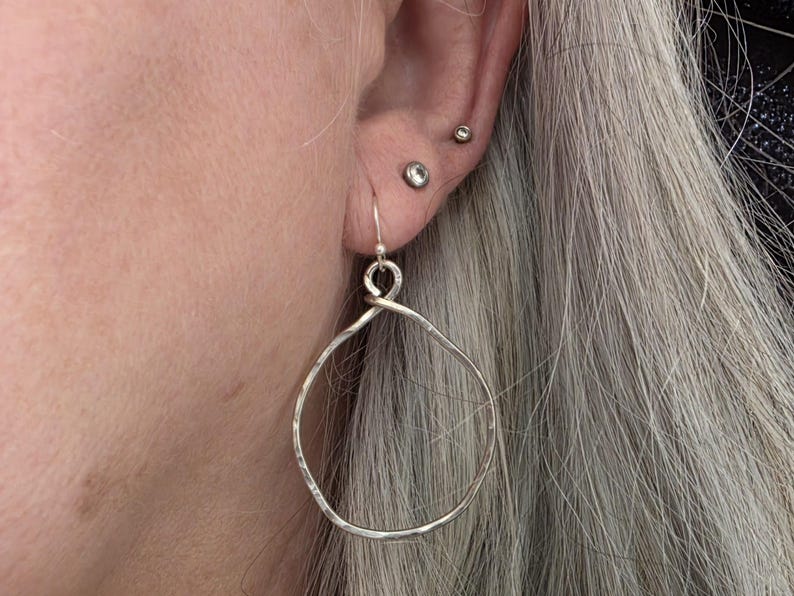 May include: Close-up of a person's ear with silver hoop earrings. The earrings are large, hammered metal hoops with a simple hook closure. The person has grey hair and multiple ear piercings.