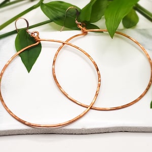 May include: Large, hammered copper hoop earrings. The earrings are round with a textured surface and are suspended from copper ear wires. The earrings are displayed on a white surface with green leaves in the background.