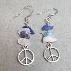 May include: A pair of dangle earrings featuring a silver peace symbol charm. Each earring has a stack of blue, white, and clear gemstone chips. The earrings have silver-colored ear wires.