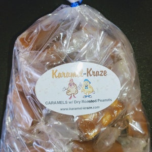 Może przedstawiać: Przezroczysty plastikowy woreczek wypełniony karmelkami z prażonymi orzeszkami ziemnymi. Woreczek ma etykietę z napisem "Karamel-Kraze" z kresk&oacute;wkowym obrazkiem dw&oacute;ch karmelk&oacute;w w nakryciach głowy z orzeszk&oacute;w ziemnych. Etykieta zawiera r&oacute;wnież napis "Caramels w/ Dry Roasted Peanuts" i "www.karamel-kraze.com".