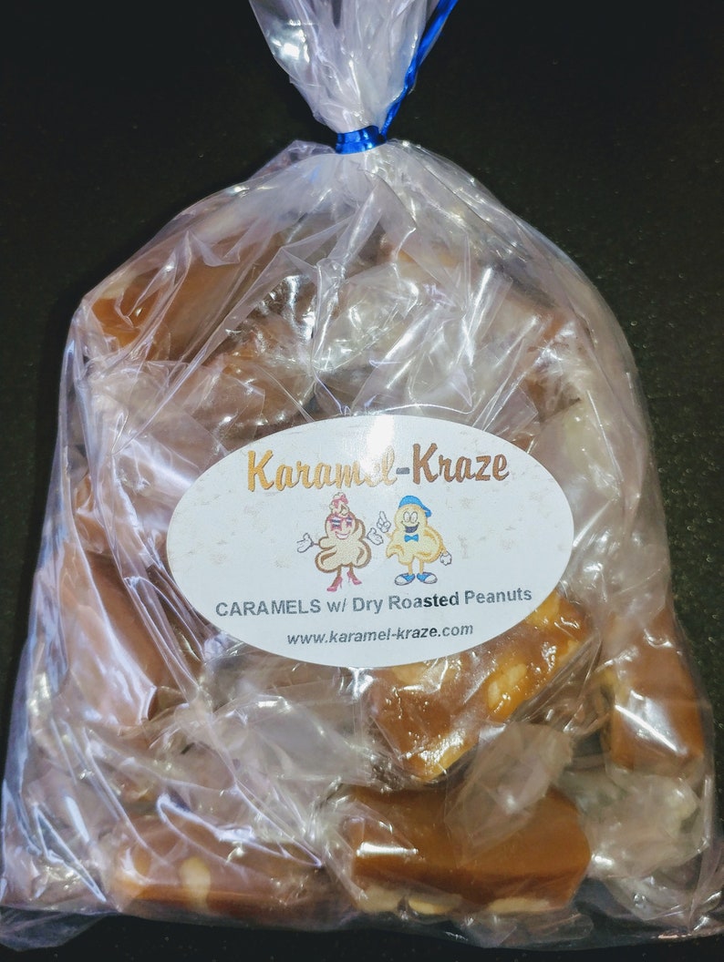 Może przedstawiać: Przezroczysty plastikowy woreczek wypełniony karmelkami z prażonymi orzeszkami ziemnymi. Woreczek ma etykietę z napisem "Karamel-Kraze" z kresk&oacute;wkowym obrazkiem dw&oacute;ch karmelk&oacute;w w nakryciach głowy z orzeszk&oacute;w ziemnych. Etykieta zawiera r&oacute;wnież napis "Caramels w/ Dry Roasted Peanuts" i "www.karamel-kraze.com".