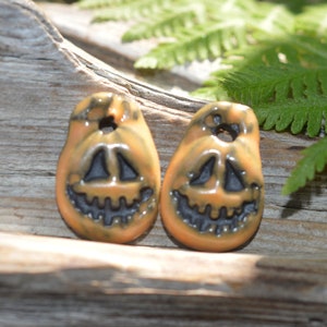 May include: Two orange ceramic pumpkin pendants with black facial features and a hole at the top for stringing. The pendants are on a weathered wooden surface with green fern leaves in the background.