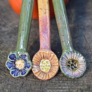 May include: Three ceramic spoons with flower-shaped bowls. The spoons are green, yellow, and orange with brown speckles. The bowls are decorated with blue, yellow, and orange flowers.