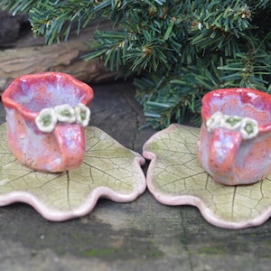 May include: Two small pink ceramic teacups with white flower accents, each sitting on a green and pink leaf-shaped saucer.