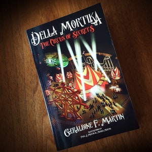 Op de afbeelding: Boekomslag voor "Della Mortika: The Circus of Secrets" van Geraldine F. Martin. De omslag toont een kleurrijke illustratie van een circustent, een reuzenrad en een menigte mensen. De titel staat in witte letters op een zwarte achtergrond.
