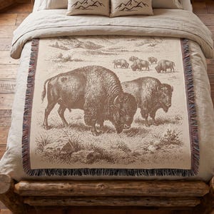 May include: A woven throw blanket featuring a brown and cream illustration of two large bison in the foreground and a herd of smaller bison in the background. The blanket has a fringed edge and is draped over a bed with matching mountain pillows.