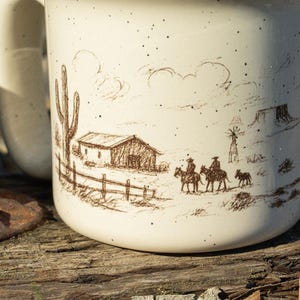 May include: Cream ceramic mug featuring a brown illustration of a desert landscape. The design includes a cactus, a barn, cowboys on horseback, and a windmill. The mug has a speckled finish and a large handle.