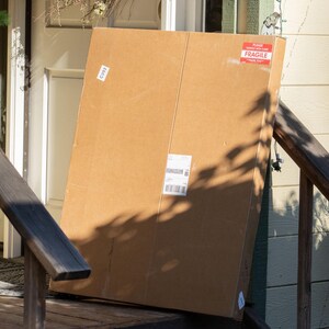May include: A large, rectangular brown cardboard shipping box standing upright. The box features a red and white rectangular sticker in the top right corner that reads "PLEASE HANDLE WITH CARE FRAGILE THANK YOU" and a white shipping label in the center.