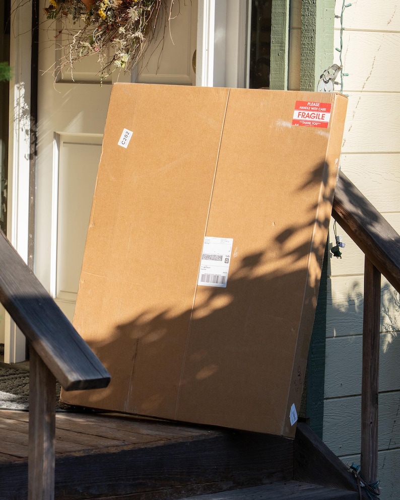 May include: A large, rectangular brown cardboard shipping box standing upright. The box features a red and white rectangular sticker in the top right corner that reads "PLEASE HANDLE WITH CARE FRAGILE THANK YOU" and a white shipping label in the center.