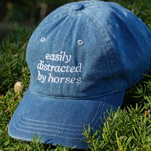 May include: A blue denim baseball cap with the embroidered text "easily distracted by horses" in white. The cap has a curved brim and a rounded crown. The hat is surrounded by green foliage.