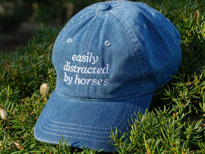 May include: A blue denim baseball cap with the embroidered text "easily distracted by horses" in white. The cap has a curved brim and a rounded crown. The hat is surrounded by green foliage.