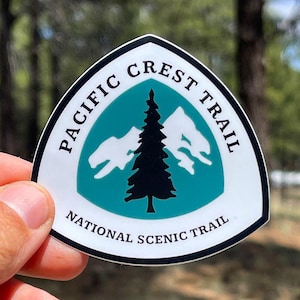 May include: A shield-shaped sticker featuring a teal center with a black pine tree silhouette and white mountain peaks. The text PACIFIC CREST TRAIL is arched at the top, and NATIONAL SCENIC TRAIL is printed at the bottom in black capital letters.