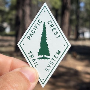 May include: A white and green diamond-shaped sticker featuring the text "PACIFIC CREST TRAIL SYSTEM" and a green tree design. The sticker is held up against a blurred outdoor background.