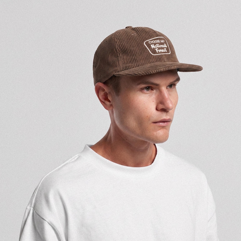 May include: A brown corduroy baseball cap featuring a white embroidered patch on the front. The patch is rectangular with rounded corners and contains the white text "CHOOSE ANY National Forest" in a stylized, cursive and sans-serif font.