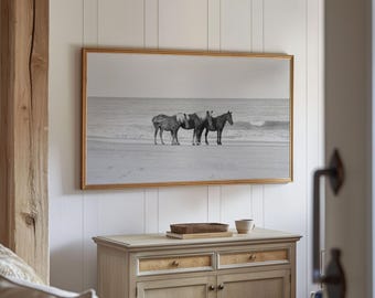 Puede incluir: Una fotografía en blanco y negro de tres caballos de pie en una playa. Los caballos están mirando hacia el océano, y las olas rompen en el fondo. La fotografía está enmarcada en un marco dorado y colgada en una pared blanca.