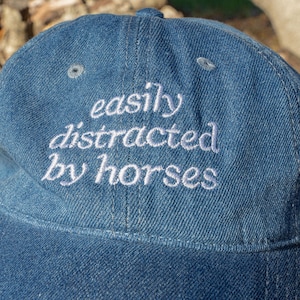 May include: A blue denim baseball cap with the embroidered phrase "easily distracted by horses" in white script. The cap has a curved brim and a button on top.