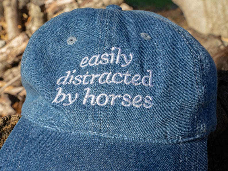May include: A blue denim baseball cap with the embroidered phrase "easily distracted by horses" in white script. The cap has a curved brim and a button on top.