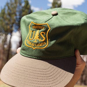 May include: A green baseball cap with a tan brim and a tan button on top. The front features an embroidered yellow shield-shaped patch with the text PROTECT PUBLIC LANDS and the letters US with a pine tree icon in the center.
