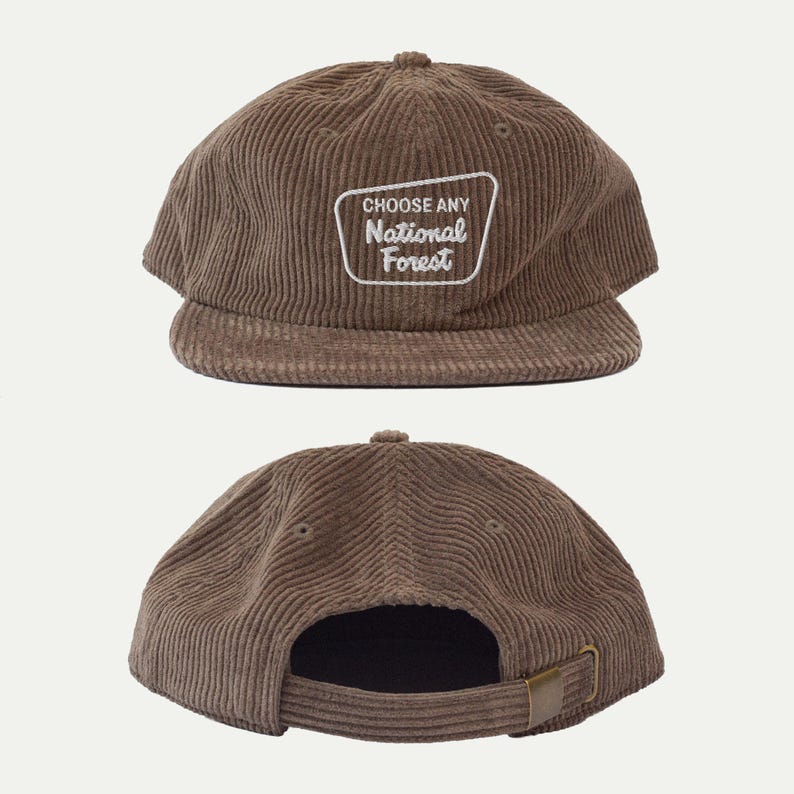May include: A brown corduroy baseball cap featuring a white embroidered patch on the front with the text CHOOSE ANY National Forest. The cap has a structured crown, a flat brim, and an adjustable strap at the back with a metal buckle closure.