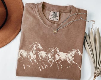 May include: A brown short-sleeve t-shirt featuring a cream-colored graphic of three horses running. The shirt has a Comfort Colors brand label at the neck with text reading S P CH. A silver-toned necklace with a small circular pendant rests on the shirt.