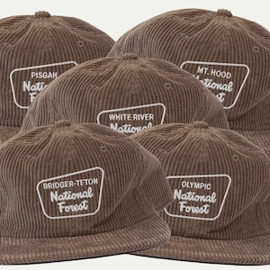 May include: A collection of brown corduroy baseball caps, each featuring a white embroidered patch with the name of a specific National Forest, including Pisgah, Mt. Hood, White River, Bridger-Teton, and Olympic, all with a structured crown.