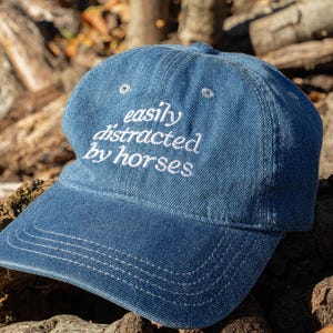 May include: A blue denim baseball cap with the embroidered text "easily distracted by horses" in white. The cap has a curved peak and a six-panel design. The hat is resting on a wooden surface.