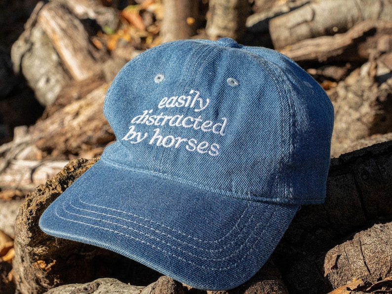May include: A blue denim baseball cap with the embroidered text "easily distracted by horses" in white. The cap has a curved peak and a six-panel design. The hat is resting on a wooden surface.