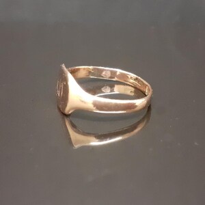 May include: A gold signet ring with a smooth, oval-shaped top. The ring is on a dark, reflective surface.