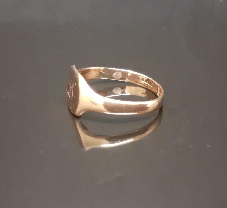 May include: A gold signet ring with a smooth, oval-shaped top. The ring is on a dark, reflective surface.