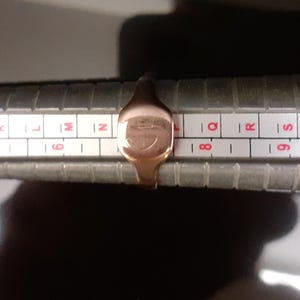 May include: A gold signet ring with a design on the top, measured on a ring sizer. The ring size is between 8 and 9.