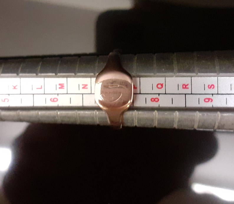 May include: A gold signet ring with a design on the top, measured on a ring sizer. The ring size is between 8 and 9.