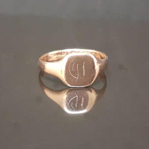 May include: A gold signet ring with a square-shaped top featuring an engraved design. The ring is made of a shiny metal and has a simple band.
