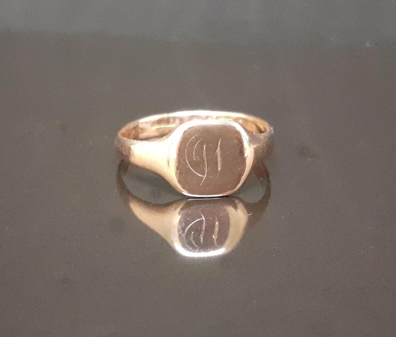May include: A gold signet ring with a square-shaped top featuring an engraved design. The ring is made of a shiny metal and has a simple band.