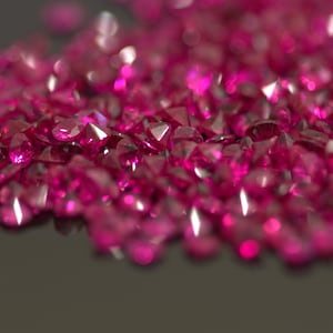 May include: A close-up of a collection of deep pink, faceted gemstones. The gems are scattered on a dark surface, reflecting light and creating bright highlights. The image evokes a sense of luxury and sparkle.