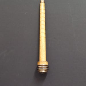 May include: A tapered wooden spindle with a light tan finish and horizontal grooves along its length. The base features a wider wooden section wrapped with two silver-coloured metal rings, and a thin metal rod extends from the top point.