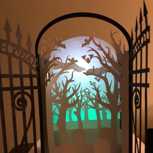 May include: A black papercraft gate opens to reveal a spooky forest scene with bats flying through the trees. The forest is illuminated with a soft blue light and the trees are silhouetted against the light. The gate is decorated with the word "Cemetery" on the top.