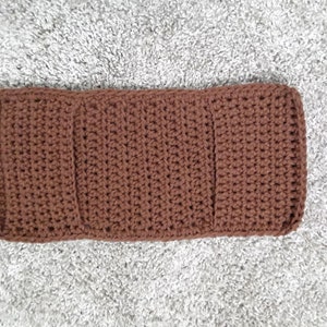 May include: A brown crocheted rectangle with three vertical sections. The crocheted rectangle is on a gray fuzzy surface.